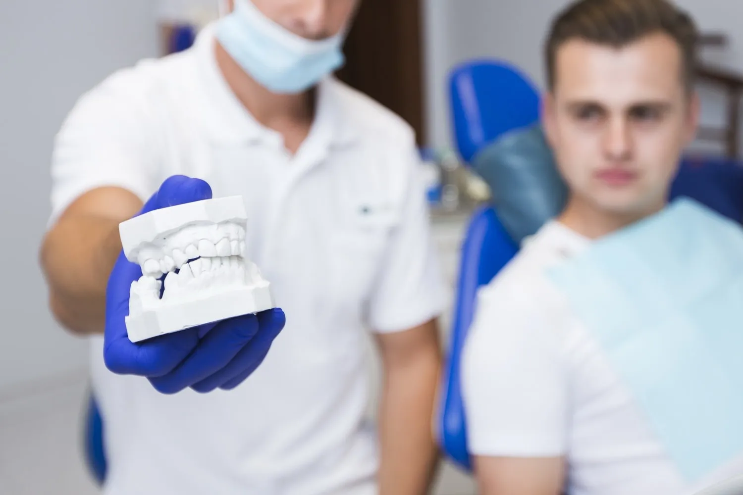 Dentista con guantes azules muestra un modelo de dentadura a un paciente sentado en el sillón de la clínica