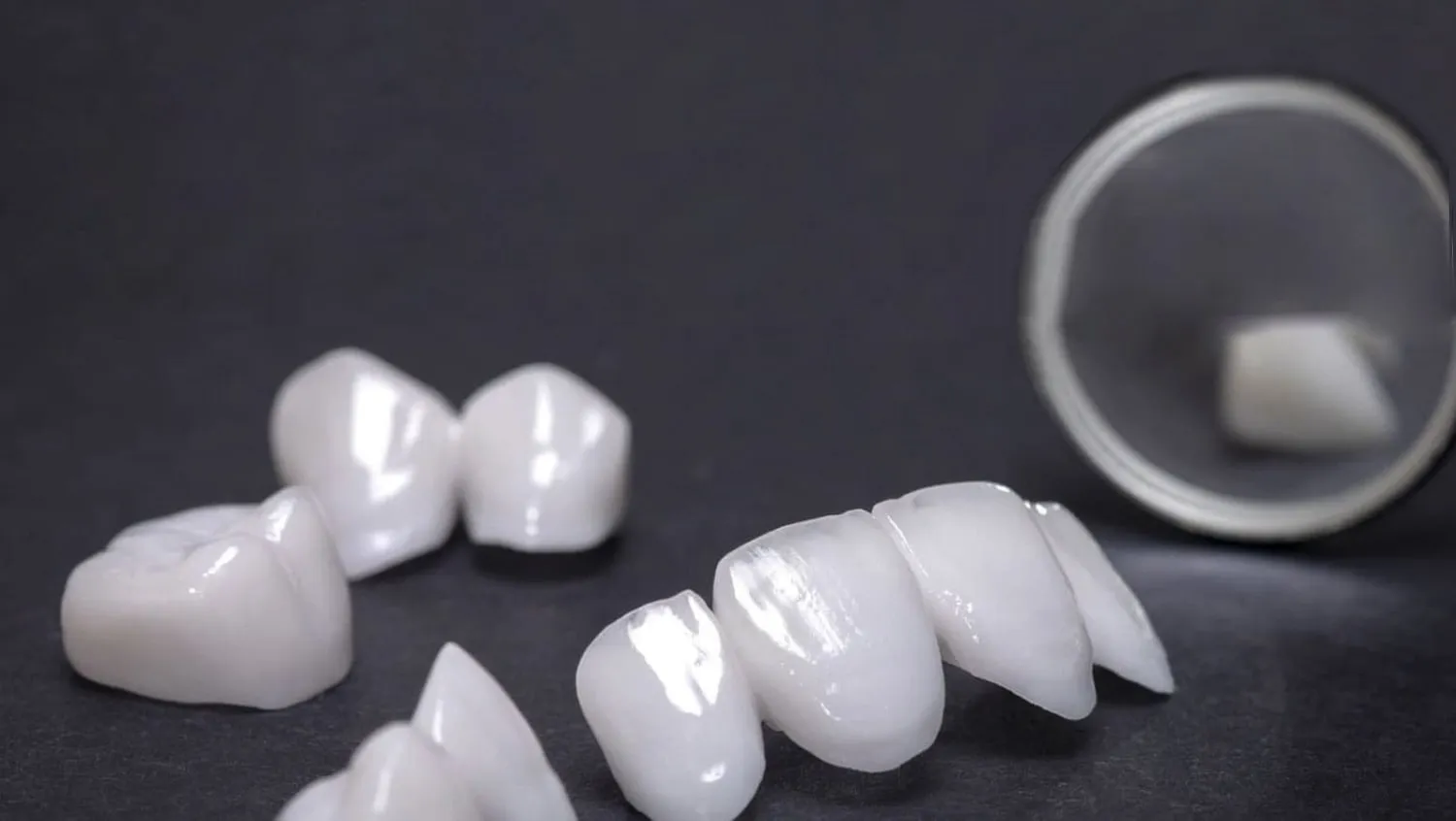 Coronas y puentes dentales blancos de porcelana sobre fondo oscuro, junto a un espejo dental
